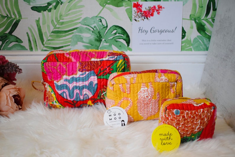 May include: Three quilted cosmetic bags with vibrant floral and abstract patterns in red, yellow, and blue. The bags are on a fluffy white surface, with a sign that says "Hey Gorgeous!" in the background.