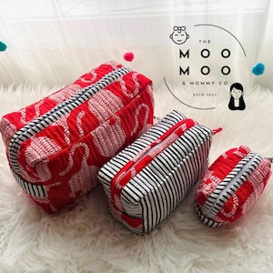 May include: Set of three red and white patterned cosmetic bags with black and white striped accents. The bags have zippers and are in varying sizes. The text "THE MOO MOO & MOMMY CO. ESTD 2013" is visible.