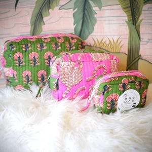May include: A set of three quilted cosmetic bags in varying sizes. The bags feature a vibrant color palette with pink and green floral and tiger print patterns. Each bag has a zipper closure and a small handle. The bags are displayed on a white, fluffy surface.