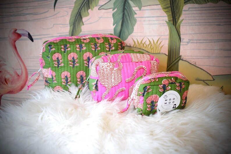 May include: A set of three quilted cosmetic bags in varying sizes. The bags feature a vibrant color palette with pink and green floral and tiger print patterns. Each bag has a zipper closure and a small handle. The bags are displayed on a white, fluffy surface.