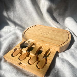 May include: A wooden cheese board with a built-in drawer that holds five cheese knives. The board is oval shaped and has a light brown colour. The knives have wooden handles and stainless steel blades. The board has the text "SnackScript" engraved on it.