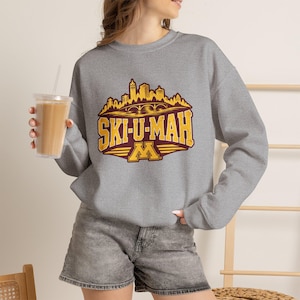 Könnte beinhalten: Ein graumeliertes Sweatshirt mit Rundhalsausschnitt und einem kastanienbraun-goldenen Grafikdruck, der eine Stadtsilhouette über dem Schriftzug "SKI-U-MAH" und einem großen "M"-Logo zeigt. Das Design weist eine abgenutzte Textur auf.
