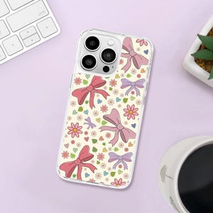 Könnte beinhalten: Transparente Handyhülle mit einem sich wiederholenden Muster aus rosa und lila Schleifen, Blumen, Herzen und Blättern auf cremefarbenem Hintergrund. Die Hülle liegt auf einer hellvioletten Oberfläche, neben einer Tastatur, einer kleinen Pflanze und einer Tasse Kaffee.