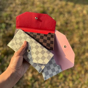 May include: Three small wallets in different colours and patterns. One is red, another is pink, and the third is a checkered pattern in black and brown. The fourth wallet is a checkered pattern in white and grey.