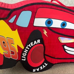 May include: A red Lightning McQueen pi&ntilde;ata with black accents, blue eyes, and a smiling mouth. The number "95" is displayed in white and orange on a yellow background. The wheels say "LIGHTYEAR".