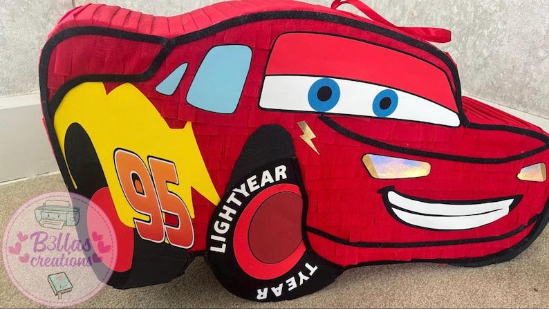 May include: A red Lightning McQueen pi&ntilde;ata with black accents, blue eyes, and a smiling mouth. The number "95" is displayed in white and orange on a yellow background. The wheels say "LIGHTYEAR".