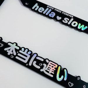 Puede incluir: Marco de matrícula negro con texto holográfico que dice "hella slow" y "DOPE VIBES FOREVER". El marco inferior tiene texto japonés y corazones y estrellas decorativos.