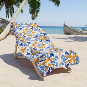 May include: A white beach towel with a blue and gold geometric pattern. The towel is draped over a wooden beach chair on a sandy beach.