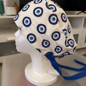 Puede incluir: Un gorro de quirófano blanco con un patrón de ojo turco azul y blanco. El gorro tiene lazos azules.