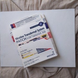 Könnte beinhalten: Eine weiße Schachtel mit blauem und rotem Text, der "Nicotine Transdermal System PATCH STOP SMOKING AID" lautet. Die Schachtel enthält 7 Pflaster und einen Verhaltensunterstützungsleitfaden. Die Schachtel befindet sich auf einer weißen Oberfläche.