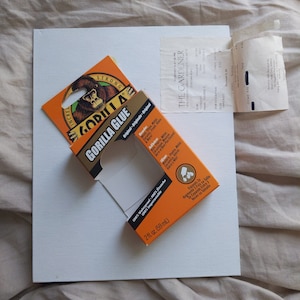 May include: A close-up of a small orange and brown box of Gorilla Glue. The box has a picture of a gorilla on it and the text "Gorilla Glue" in white letters. The box is sitting on a white surface.