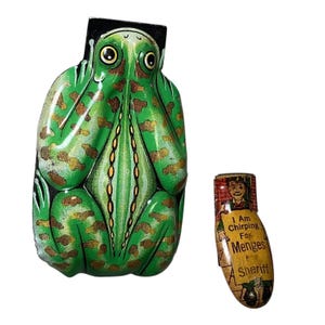 Pode incluir: Sapo de brinquedo de lata vintage com manchas verdes e castanhas, e um pequeno brinquedo de lata com o texto "I Am Chirping For Menges For Sheriff". O sapo tem olhos grandes e amarelos e está sentado.