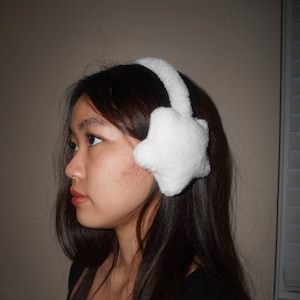 Puede incluir: Una mujer con el pelo largo y oscuro lleva orejeras blancas y peludas con forma de estrella.