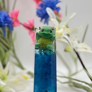 Peut inclure: Objet rectangulaire transparent en résine avec une figurine de tortue verte à l'intérieur. La moitié inférieure est bleu foncé avec des paillettes, et la tortue est au-dessus. Fleurs floues en arrière-plan.