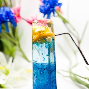 May include: A clear glass cylinder filled with blue resin and yellow and white resin. The resin has a glittery texture.