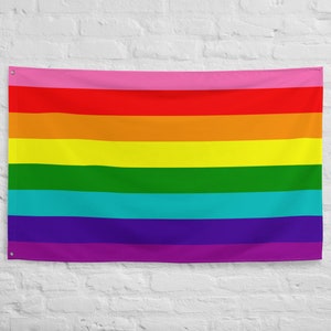 May include: A rainbow pride flag with pink, red, orange, yellow, green, turquoise, blue, and purple stripes. The flag is hanging on a white brick wall.