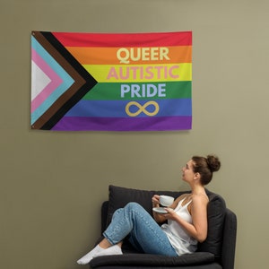 Może przedstawiać: Tęczowa flaga dumy z napisem "Queer Autistic Pride" i symbolem nieskończoności. Flaga wisi na ścianie za osobą siedzącą na kanapie.
