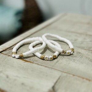 Peut inclure: Trois bracelets en perles blanches avec des breloques en lettres dorées qui épellent les lettres "CMK".