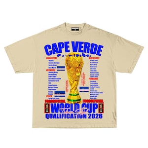 May include: Cream-colored t-shirt featuring the text "CAPE VERDE" in blue, with a golden trophy graphic. The shirt lists team members and "WORLD CUP QUALIFICATION 2026" in blue and red. The shirt has a crew neck and short sleeves.