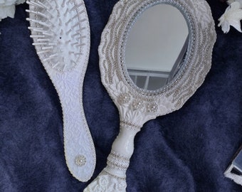 May include: A white hand mirror and hairbrush set with pearl accents and floral designs. The mirror has an oval-shaped glass and a decorative handle. The brush has a white plastic handle with a white bristle head.