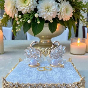 以下が含まれることがあります： ライトブルーのベルベットの結婚指輪枕。金色のトリムとクリスタルアクセントが施されています。クリスタルの羽とハートのディテールが付いた2羽の金の白鳥が上にあります。2つの金の指輪が枕の上に置かれ、結婚式に備えています。