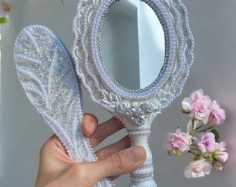 May include: A white hand mirror with a pearl and floral design. The mirror has an oval shape and is surrounded by a decorative frame. The handle is also decorated with pearls and flowers.