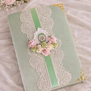 May include: A sage green journal with a delicate lace design and a green ribbon down the centre. The journal is adorned with pink and cream-coloured flowers and gold corner accents. A beautiful keepsake.