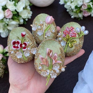 May include: Four green velvet Easter eggs decorated with different floral embellishments. Each egg features a different flower design with pink, red, and white accents. The eggs are adorned with pearls and rhinestones.