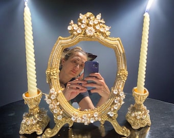 May include: A gold-toned ornate mirror with a beveled oval glass and a decorative crown-like top. The mirror is surrounded by rhinestones and sits on a stand with two candle holders on either side. The candles are white and lit.