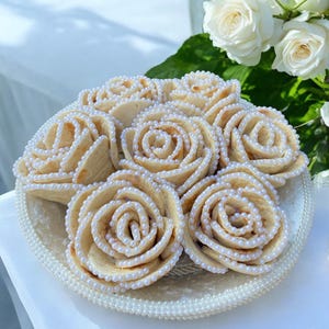 May include: A plate of rose-shaped cookies with pearl-like decorations. The cookies are light brown and arranged in a circular pattern on a decorative plate. White roses and greenery are in the background.