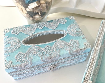 May include: A light blue tissue box cover with a silver lace design and pearl accents. The cover has a rectangular shape and a tissue opening on the top.