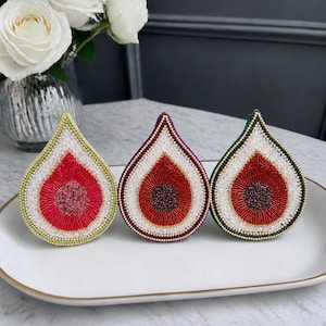 Peut inclure: Trois broches en forme de figue, ornées de perles rouges, blanches et vertes, présentées sur une assiette ovale blanche. Chaque broche présente un motif central rouge, entouré de perles blanches et d'une bordure colorée. Les broches mesurent environ 5 cm de long.