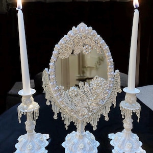 May include: A white vanity mirror set with two candle holders. The mirror and candle holders are decorated with pearl-like beads and rhinestones. The candles are white and lit.