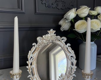 May include: An oval tabletop mirror with an ornate white and gold frame decorated with floral accents and crystals, flanked by two matching candle holders, each holding a tall, slender white taper candle, all featuring intricate detailing.