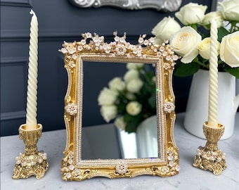 May include: A gold-framed mirror with ornate floral and crystal embellishments. Two gold candle holders with cream-colored candles flank the mirror. A vase of white roses is in the background, creating an elegant, decorative display.