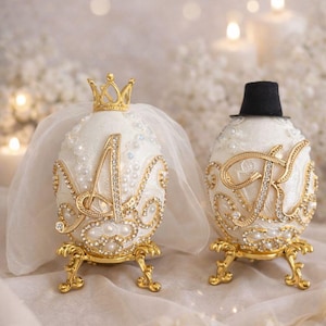 May include: Two ornate, white egg-shaped decorative items, one with a gold crown and veil, the other with a black top hat. Both are embellished with gold filigree, rhinestones, and pearls, and sit on gold stands. The letter "A" and "K" are displayed on the eggs.