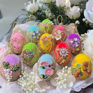 May include: A collection of ten colourful, intricately decorated Easter eggs. Each egg is made of velvet and adorned with beads, sequins, and floral embellishments. The eggs are arranged in a basket with white flowers.