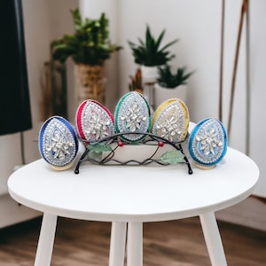 May include: Five decorative eggs with intricate designs and rhinestones, displayed on a black metal stand. The eggs are blue, red, green, yellow, and light blue.