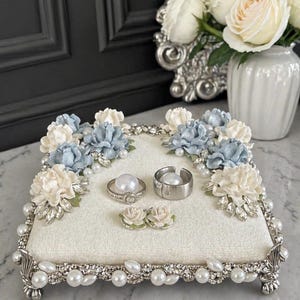 May include: A square, cream-coloured ring holder with a pearl and rhinestone border. The holder is decorated with blue and white fabric flowers and holds three rings and two small floral accents. A vase of white roses sits in the background.