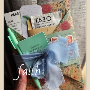May include: A gift wrapped in brown paper with a floral pattern. The gift is tied with a light blue ribbon and includes a small green notebook with the text "The Lord has done great" on the cover, a tea bag with the text "TAZO Regenerative Organic Awake", and a green pen.