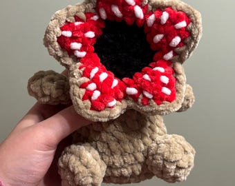 May include: A crocheted Demogorgon plushie in tan, red, white, and black. The plushie has a flower-like head with red petals dotted with white spots, a black center, and a tan body. A unique handmade item.