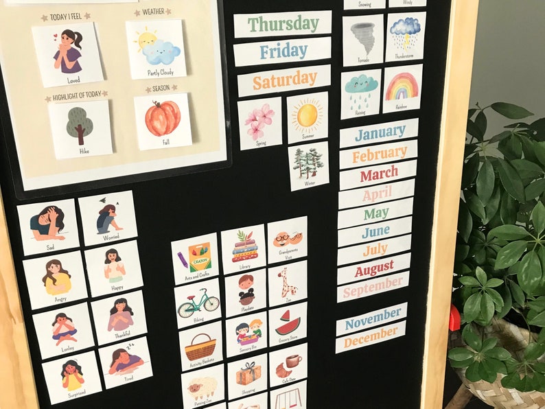 May include: A black dry-erase board with colourful magnetic cards. The cards feature images and text related to days of the week, weather, seasons, emotions, and months of the year. The text on the cards includes "Thursday", "Friday", "Saturday", "January", "February", "March", "April", "May", "June", "July", "August", "September", "November", "December", "Today I Feel", "Weather", "Partly Cloudy", "Season", "Highlight of Today", "Loved", "Tree", "Sad", "Worried", "Angry", "Happy", "Lonely", "Thankful", "Surprised", "Tired", "Arts and Crafts", "Library", "Playtime", "Swing", "Water", "Pumpkin", "Fall", "Spring", "Summer", "Snowing", "Wind", "Thunderstorm", "Rainy", "Rainbow".