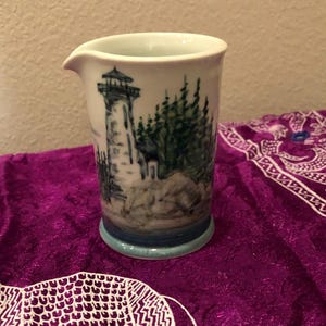 May include: A white ceramic creamer with a spout, featuring a hand-painted scene of a lighthouse, trees, and rocks in shades of blue and green. The creamer has a light blue base.