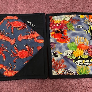 May include: Two blue fabric pot holders with a nautical theme. One features red lobsters and crabs on a blue background with the word "MAINE" embroidered on the corner. The other features colorful fish, coral, and sea life on a blue background.