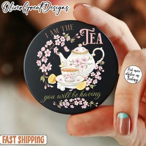 Puede incluir: Soporte redondo negro para teléfono con un diseño floral de té. El diseño presenta una tetera, una taza de té, limones y el texto "I AM THE TEA you will be having".
