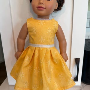 May include: A doll with dark brown hair and brown eyes wearing a sleeveless yellow dress featuring a white starburst pattern and a thin white ribbon waistband. The dress has a pleated skirt that flares out from the waistline to the hem.