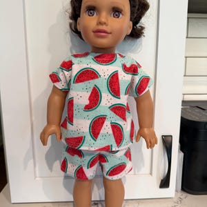 May include: A doll with brown hair and brown eyes wearing a short-sleeved shirt and matching shorts. The clothing features a repeating pattern of red watermelon slices on a light mint green background with thin white horizontal stripes.