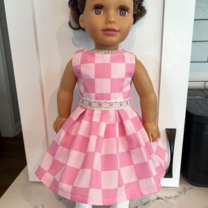 May include: A doll wearing a pink and white checkered dress with a floral belt and white shoes. The doll has brown hair and is standing in front of a white door.