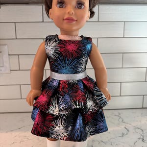 May include: A doll wearing a black dress with a colorful firework pattern in red, white, and blue. The dress has a silver belt and a tiered skirt. The doll is also wearing white boots.