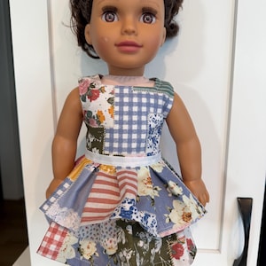 May include: A doll wearing a patchwork dress with floral, gingham, and striped patterns. The dress has a white ribbon belt and a layered skirt. The doll has brown hair, brown eyes, and is wearing white boots.